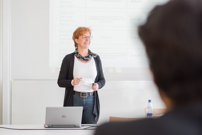 Eine Lehrperson steht vor einer Beamerprojektion und erklärt etwas.